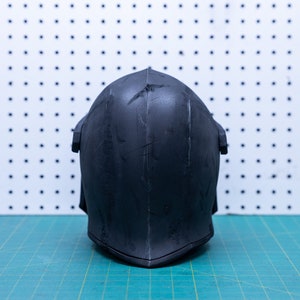 Op de afbeelding: Een zwarte helm met een puntige bovenkant en een V-vormige onderkant. De helm heeft verticale lijnen en een paar krassen. De achtergrond is een wit pegboard met zwarte stippen en een groene snijmat.