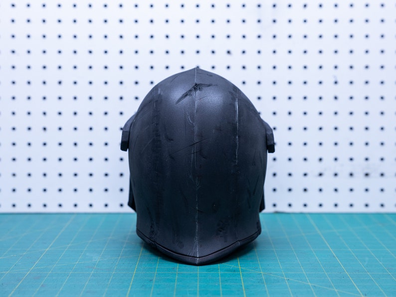 Op de afbeelding: Een zwarte helm met een puntige bovenkant en een V-vormige onderkant. De helm heeft verticale lijnen en een paar krassen. De achtergrond is een wit pegboard met zwarte stippen en een groene snijmat.