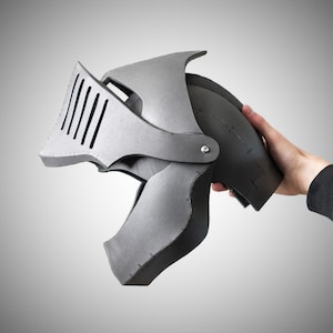 May include: A grey knight helmet constructed from foam panels with visible seams and a hinged visor featuring vertical slits. The helmet has a rounded dome shape and a pointed front section, showcasing a matte finish and a metallic appearance.