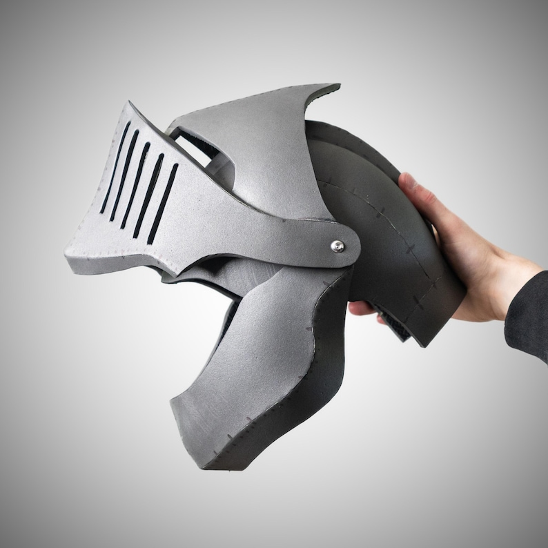 May include: A grey knight helmet constructed from foam panels with visible seams and a hinged visor featuring vertical slits. The helmet has a rounded dome shape and a pointed front section, showcasing a matte finish and a metallic appearance.
