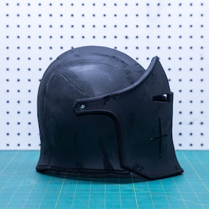 Op de afbeelding: Een zwarte middeleeuwse helm met vizier en kruisontwerp. De helm is gemaakt van donker materiaal en staat op een groen en wit oppervlak. De helm heeft een afgeronde bovenkant en een gedetailleerd vizier.