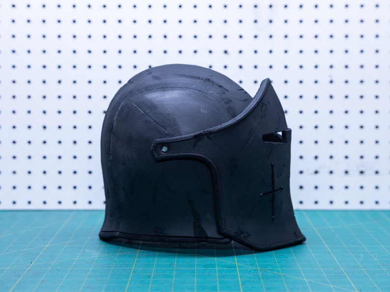 Op de afbeelding: Een zwarte middeleeuwse helm met vizier en kruisontwerp. De helm is gemaakt van donker materiaal en staat op een groen en wit oppervlak. De helm heeft een afgeronde bovenkant en een gedetailleerd vizier.