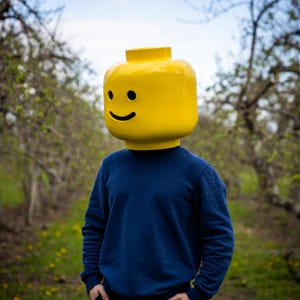 May include: A person wearing a large yellow Lego head with a smiling face, paired with a navy blue sweatshirt. The background features a blurred outdoor scene with trees and greenery, creating a playful and whimsical aesthetic.