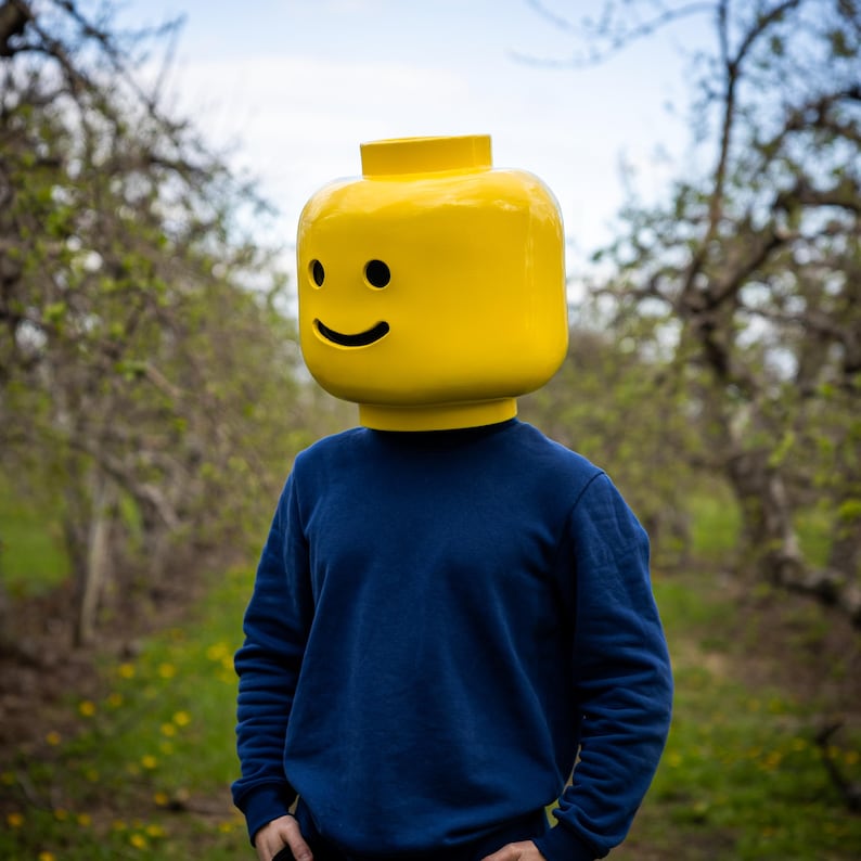 May include: A person wearing a large yellow Lego head with a smiling face, paired with a navy blue sweatshirt. The background features a blurred outdoor scene with trees and greenery, creating a playful and whimsical aesthetic.