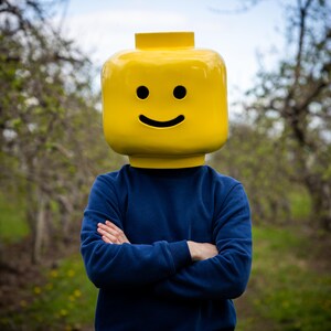 May include: A person wearing a large, yellow Lego head with a smiling face. The person is wearing a navy blue sweatshirt, arms crossed. The background is a blurred outdoor scene with trees and greenery.