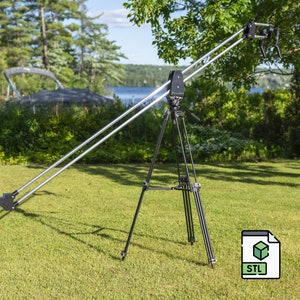 Puede incluir: Una grúa de cámara negra con un brazo de pluma plateado, montada en un trípode negro, sobre un césped verde. La grúa está extendida y el brazo de pluma apunta hacia la derecha. La grúa se encuentra en un entorno de parque con árboles y un lago en el fondo. Archivo STL.