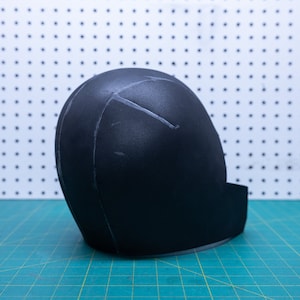 May include: A black foam helmet with white lines drawn on it. The helmet is sitting on a green cutting mat.