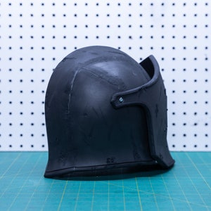 Op de afbeelding: Een zwarte helm met vizier, waarschijnlijk voor historische re-enactment of kostuumgebruik. De helm is gemaakt van een donker materiaal en ligt op een groene snijmat. De achtergrond is een wit pegboard.