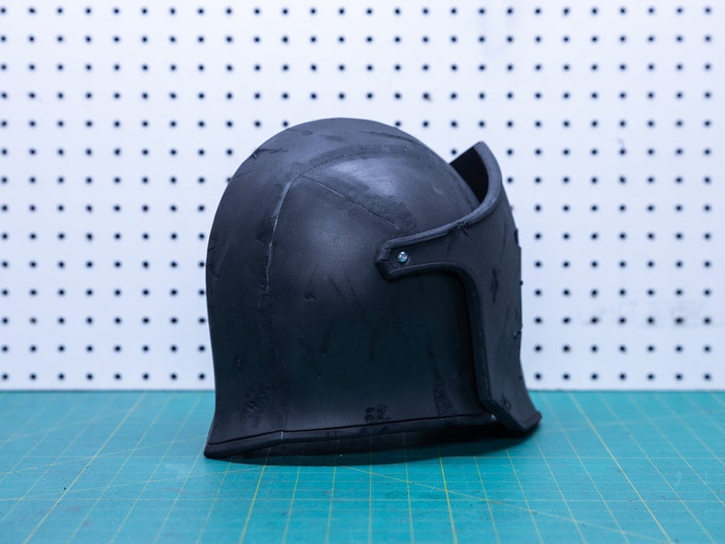 Op de afbeelding: Een zwarte helm met vizier, waarschijnlijk voor historische re-enactment of kostuumgebruik. De helm is gemaakt van een donker materiaal en ligt op een groene snijmat. De achtergrond is een wit pegboard.