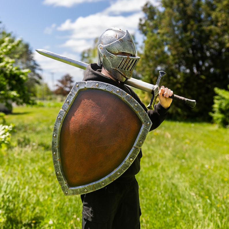 May include: A silver knight helmet with a visor, a brown heater shield with a silver-toned riveted border, and a long silver sword held across the shoulders. The gear features a weathered, metallic finish and is worn over a black hooded garment.