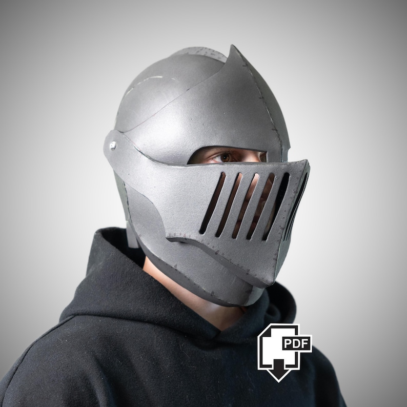 May include: A silver-colored knight helmet featuring a rounded dome, a pointed visor, and a face guard with five vertical ventilation slits. The helmet is constructed from multiple joined sections and is worn over a black hooded sweatshirt.