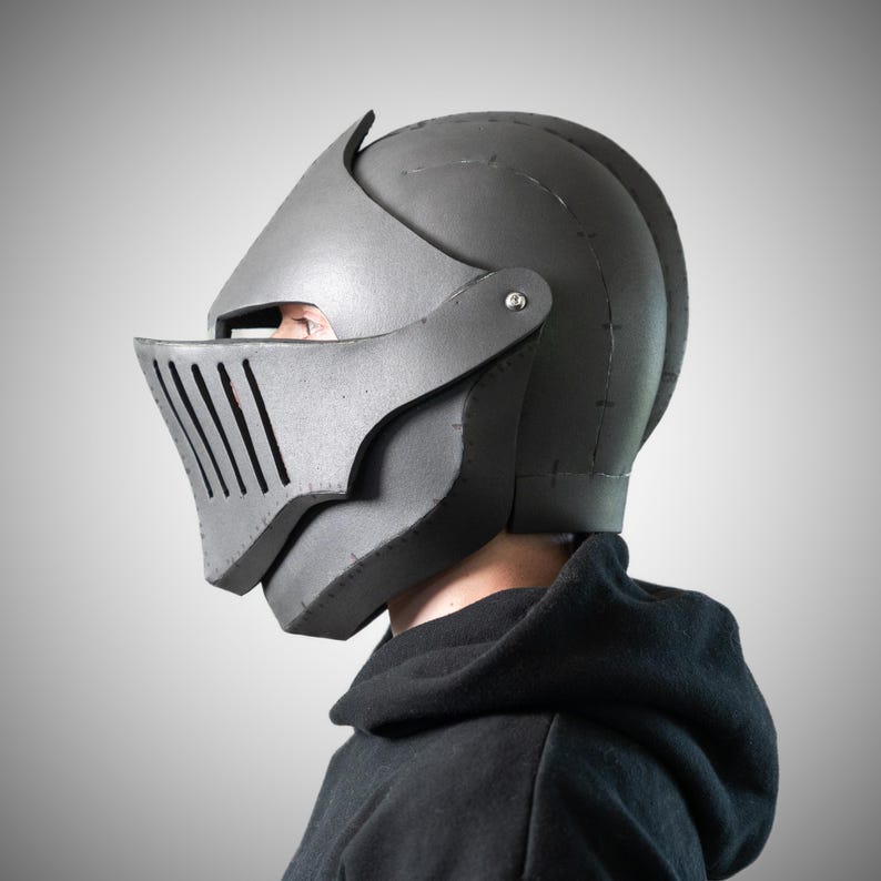 May include: A matte dark gray knight helmet featuring a pointed visor with vertical ventilation slits, a rounded dome structure, and visible seam lines along the side panels, secured with a metallic rivet at the temple area.