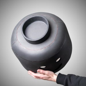 May include: A large, black, Lego-style head with a smiling face and a circular opening on top. The head is held in a person's hand against a grey background. The head has a matte finish and visible seams.