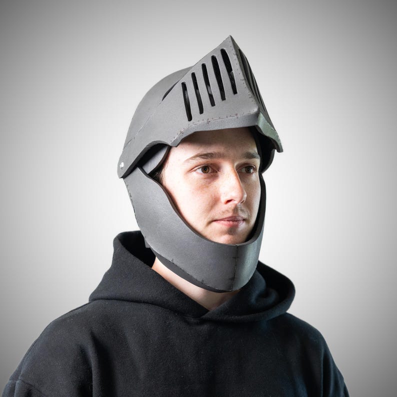 May include: A grey knight helmet featuring a pointed visor with vertical ventilation slits and a chin guard. The helmet is constructed from a smooth, matte material and covers the upper head and jaw area, leaving the face partially visible.