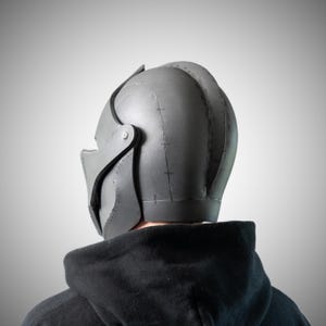May include: A matte gray helmet featuring a rounded dome shape with visible seam lines and dark stitching details along the construction points. The helmet includes a hinged side panel secured with a silver-toned metal screw and a visor section.
