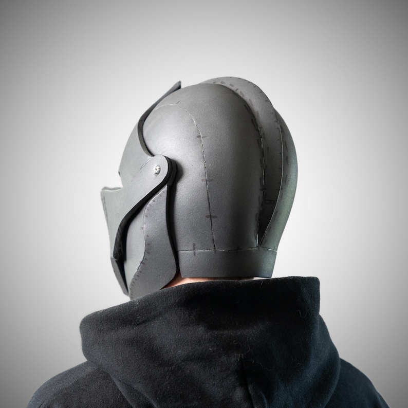 May include: A matte gray helmet featuring a rounded dome shape with visible seam lines and dark stitching details along the construction points. The helmet includes a hinged side panel secured with a silver-toned metal screw and a visor section.