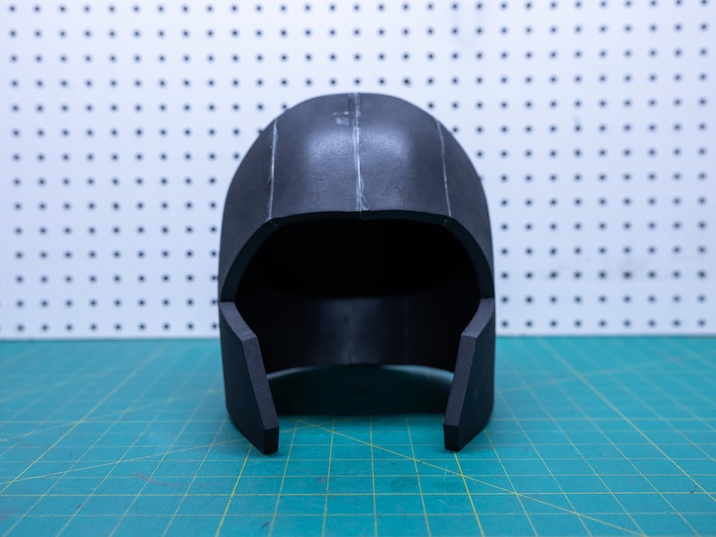 May include: A black foam helmet with a smooth, rounded top and a slightly pointed chin guard. The helmet is sitting on a green cutting mat.