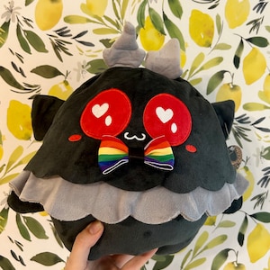 以下が含まれることがあります： 黒のぬいぐるみで、下部にグレーのフリルが付いています。おもちゃは、白いハートが入った大きな赤い目、虹色の蝶ネクタイ、頭の上に小さなグレーの角が付いています。