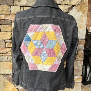 May include: A black denim jacket with a patchwork quilt design on the back. The quilt is made of pink, yellow, and blue fabric.