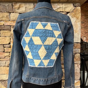 May include: A blue denim jacket with a hexagonal patchwork design on the back. The patchwork is made of blue and cream fabric and is sewn in a geometric pattern.