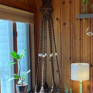 May include: A brown macrame plant hanger with a woven design and white beads hanging from the top. The hanger is suspended from the ceiling in a room with wood paneling and a window.