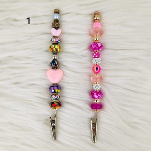 May include: Two pink and multi-colored beaded hair clips with a silver clip at the bottom. The beads are various shapes and sizes, including hearts, circles, and squares. The beads are pink, purple, blue, green, yellow, and black.