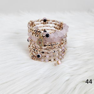 May include: Multi-strand bracelet with pink and gold beads, evil eye charms, and a heart charm. The bracelet is wrapped around itself and has a layered look.