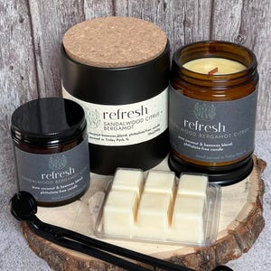 Puede incluir: Tres tarros de velas de vidrio negro y marrón con tapas de corcho. Las velas están etiquetadas con "refresh" y tienen el aroma "sandalwood bergamot citrus". Las velas están sobre una superficie de madera con una bandeja de plástico transparente de seis fundidos de cera blanca.