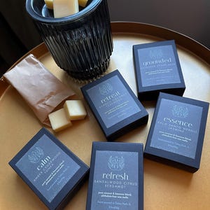 Puede incluir: Seis cajas negras con etiquetas blancas que contienen fundidos de cera perfumados. Las etiquetas tienen los nombres "Calm", "Retreat", "Grounded", "Essence", "Refresh" y "Calm". Los fundidos de cera están disponibles en una variedad de aromas, incluyendo madera de cedro ámbar, cítricos de sándalo, salvia de cedro lavanda, neroli de palo santo y teca de coco.