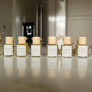 May include: Eight small glass bottles with wooden lids and reeds, each labelled with a different name. The bottles contain a golden liquid and are arranged in a row. The labels include names like "Harvest Moon" and "Grounded". The bottles are on a reflective surface.