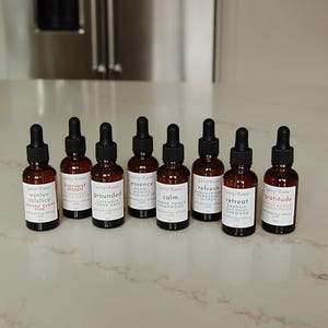 May include: Eight amber glass bottles with black droppers, each labelled with a different scent name. The bottles are arranged in a row on a white countertop. The labels include names like "Winter Solstice" and "Gratitude".