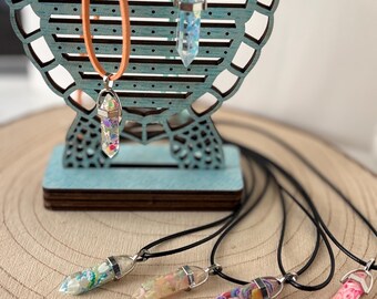 Puede incluir: Collares de punta de cristal en varios colores, exhibidos en un soporte de madera turquesa. Los colgantes están llenos de purpurina y resina de colores, suspendidos de cordones negros, naranjas y turquesas. Los collares se exhiben sobre una superficie de madera.