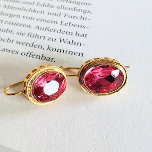 Könnte beinhalten: Ein Paar goldfarbene Ohrringe mit großen, ovalen, facettierten rosa Edelsteinen. Die Ohrringe sind in einem dekorativen Goldrahmen gefasst und liegen auf einem Buch.