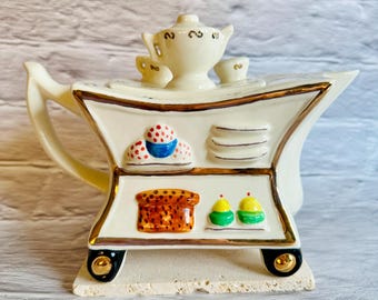Könnte beinhalten: Eine cremefarbene Keramik-Teekanne mit integriertem Servierwagen-Design. Der Wagen hat Regale mit Miniatur-Lebensmitteln, darunter Cupcakes und Toast. Goldfarbene Verzierungen akzentuieren die Teekanne und den Wagen, mit schwarzen Rädern an der Basis.
