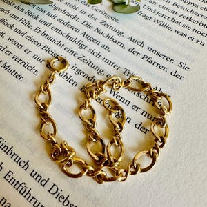 Könnte beinhalten: Ein goldfarbenes Kettenarmband mit ovalen Gliedern, das auf einer Vintage-Buchseite liegt. Das Armband besteht aus glänzendem Metall. Der Hintergrund enthält Text und kleine grüne Blätter. Das Design ist klassisch.