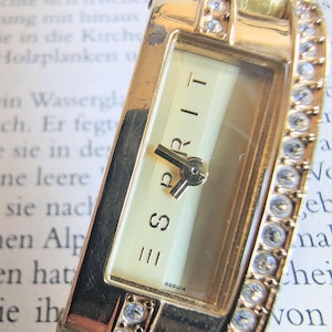 Può includere: Un orologio Esprit color oro con quadrante rettangolare e cinturino impreziosito da cristalli trasparenti. Il quadrante è color crema con lancette nere e il marchio "ESPRIT". L'orologio ha un design classico ed elegante.