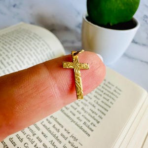 May include: A small, gold-colored cross pendant with an ornate, engraved design. The cross is held up against a blurred background of a book and a green plant in a white pot. The pendant is likely made of gold.