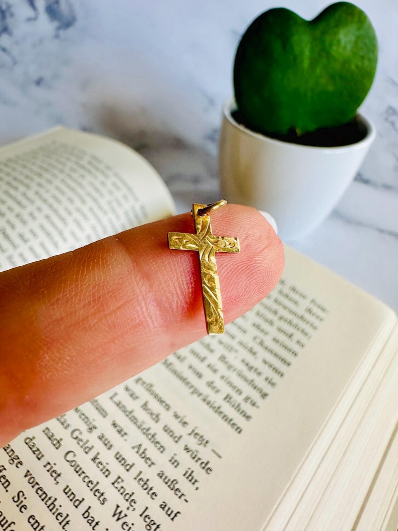 May include: A small, gold-colored cross pendant with an ornate, engraved design. The cross is held up against a blurred background of a book and a green plant in a white pot. The pendant is likely made of gold.