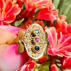 Könnte beinhalten: Ein goldener Ring mit einem einzigartigen Design, das drei Edelsteine ​​aufweist: einen grünen Smaragd, einen roten Rubin und einen blauen Saphir. Der Ring ist mit kleinen Diamanten um die Edelsteine ​​besetzt.
