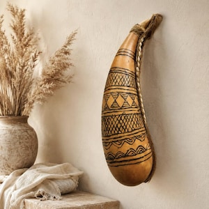 May include: A tan, teardrop-shaped gourd wall hanging featuring intricate black hand-drawn geometric patterns, including cross-hatching, diamonds, and wavy lines, accented with a thick, textured natural fibre rope and small white shell trim.