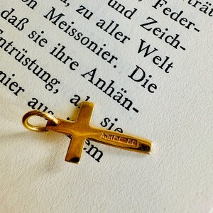 May include: A small, gold-colored cross pendant with a loop for a chain. The cross is lying on a page of a book with German text. The cross has the text "AJHOO em" engraved on it.