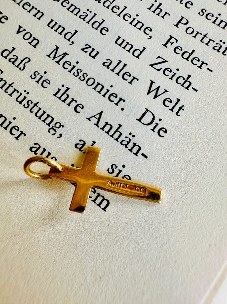 May include: A small, gold-colored cross pendant with a loop for a chain. The cross is lying on a page of a book with German text. The cross has the text "AJHOO em" engraved on it.