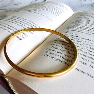 Può includere: Un braccialetto dorato è appoggiato su un libro aperto. Il braccialetto ha un design circolare semplice, che riflette la luce. Le pagine del libro sono ingiallite, con testo visibile. Lo sfondo presenta una superficie simile al marmo.