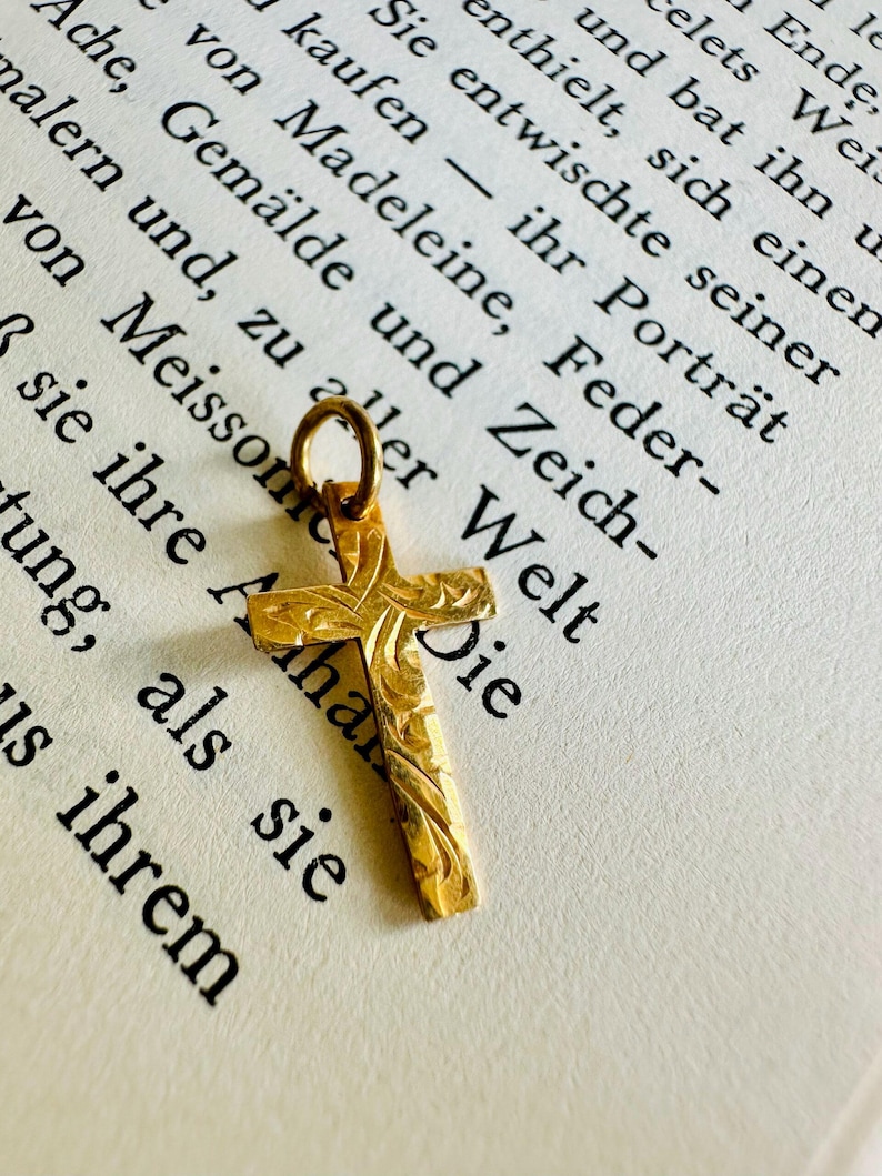 May include: A small, gold-colored cross pendant with intricate floral carvings. The cross is suspended from a small loop and rests on a page of a book with German text. The pendant is a religious symbol.