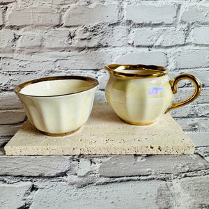 Puede incluir: Un juego de jarra de crema y azucarero de cerámica blanca con ribetes dorados. La jarra tiene un diseño acanalado y un asa. El azucarero tiene un diseño acanalado y una base plana. El juego está sobre una superficie de piedra beige claro.