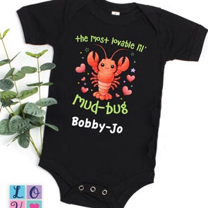 May include: Black baby bodysuit with the text "the most lovable lil' mud-bug Bobby-Jo" in green and white. A cartoon lobster with pink hearts and stars is printed on the front.