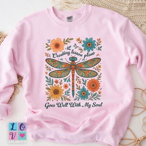 Può includere: Felpa rosa chiaro con un design di libellula colorata circondata da fiori. Il testo sulla felpa recita "Creating lesson plans Goes Well With My Soul". La felpa ha anche il testo "LOV Unlimited".