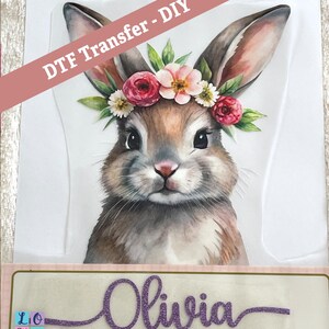May include: A watercolour illustration of a brown bunny wearing a flower crown. The bunny is looking directly at the viewer. The text "Olivia" is written in purple glitter below the bunny.