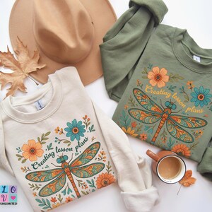 May include: Two sweatshirts, one cream and one olive green, feature a dragonfly design with the text "Creating lesson plans." The dragonfly is surrounded by flowers in orange, teal, and blue. A brown hat and a fall leaf are also in the image.