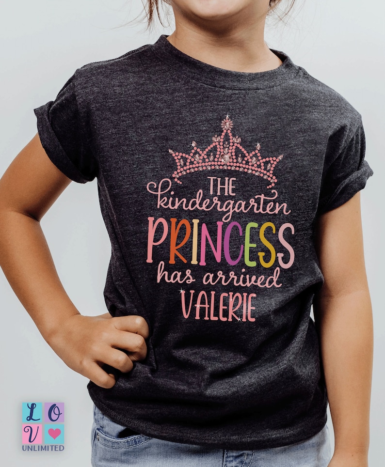 May include: A dark grey t-shirt with a pink crown and the text "The kindergarten princess has arrived Valerie". The text "princess" is in rainbow colours.